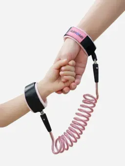 Pink stretchable safety cord connecting an adult's wrist to a child's wrist while they hold hands, with AINOMI branding visible