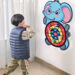 A young boy playing indoors by throwing a sticky ball at the elephant dartboard hanging on the wall